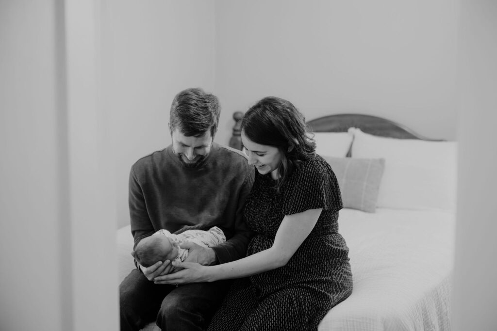 Black and white picture of parents smiling down at their baby that they're holding together at the end of their bed.