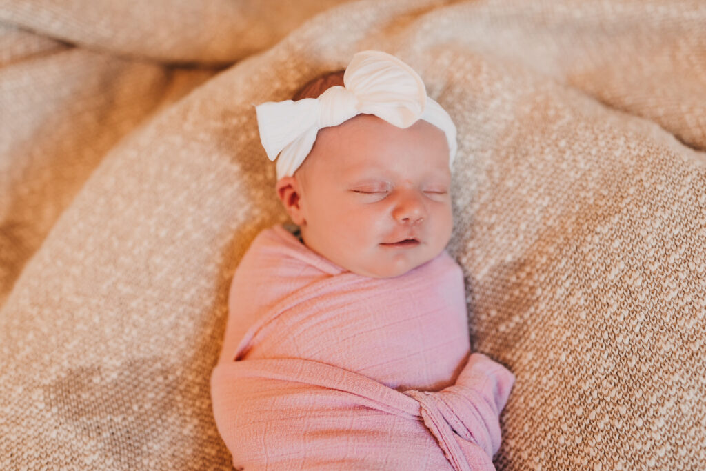 Newborn baby girl smiling while in a pink swaddle and wearing a white bow.