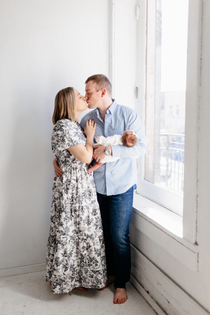 Dad holding newborn baby in one hand and holding his wife's waist in the other pulling her in for a kiss
