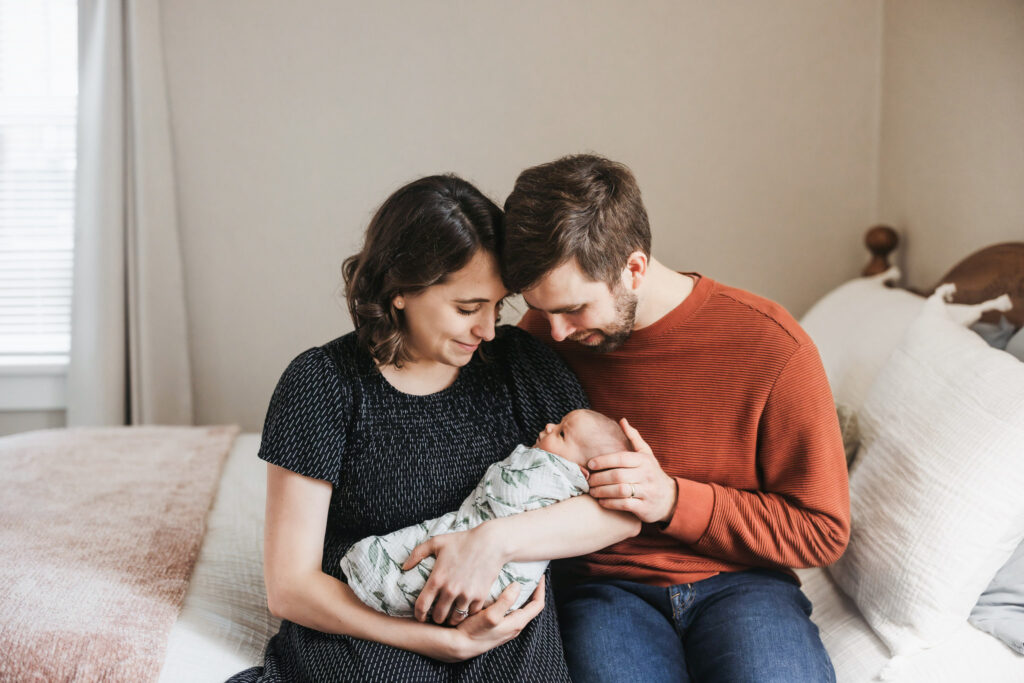 Mom holding newborn baby while her husband rubs the newborns head. The couple is sitting at the end of the their master bed.