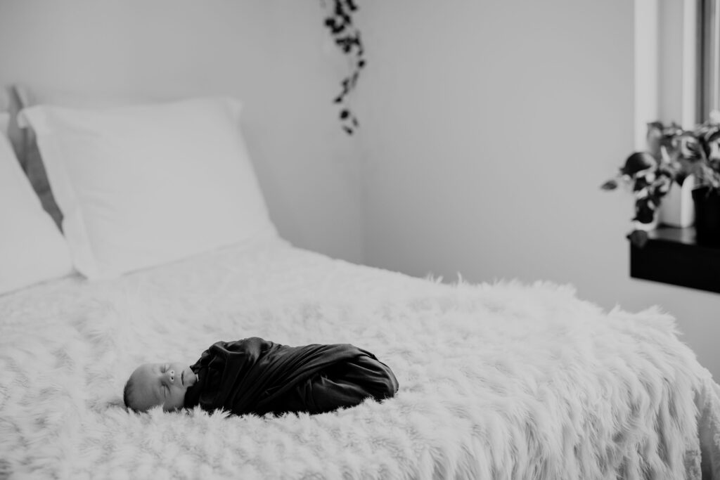 Black and white photo of newborn baby swaddled and sleeping at the end of the parents bed